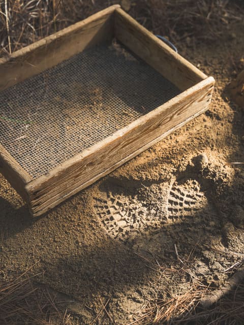 A wooden soil screen resting on freshly sifted dirt, a boot print pressed into the pile beside it.